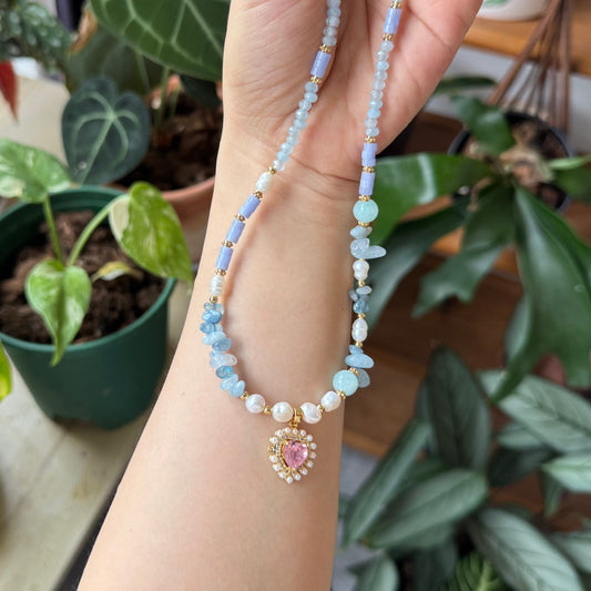 Hand holding a beaded necklace with a pink pendant in front of potted plants